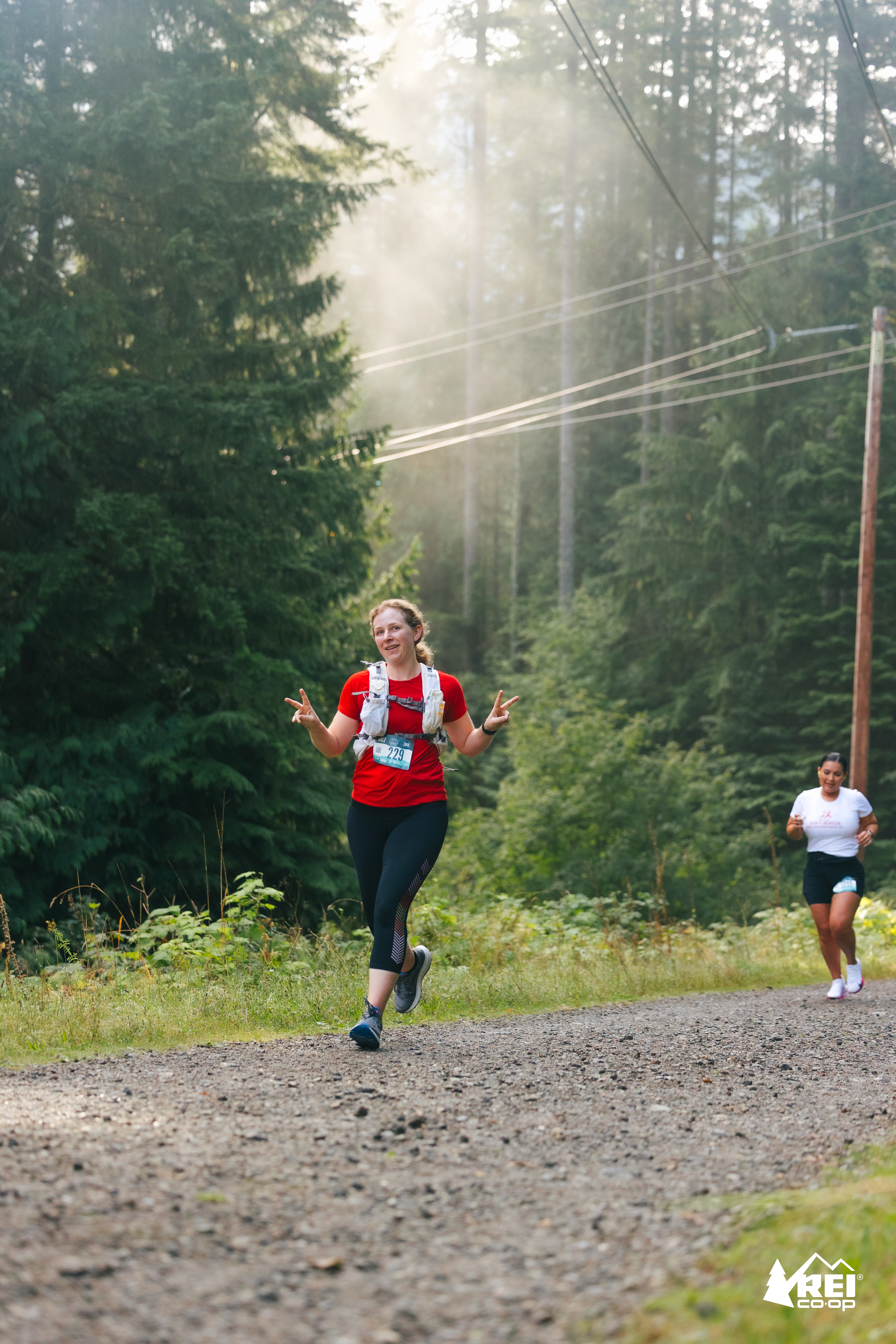 Sarah running down a gravel path in a sunlit forest, she has a relaxed smile and is flashing peace signs